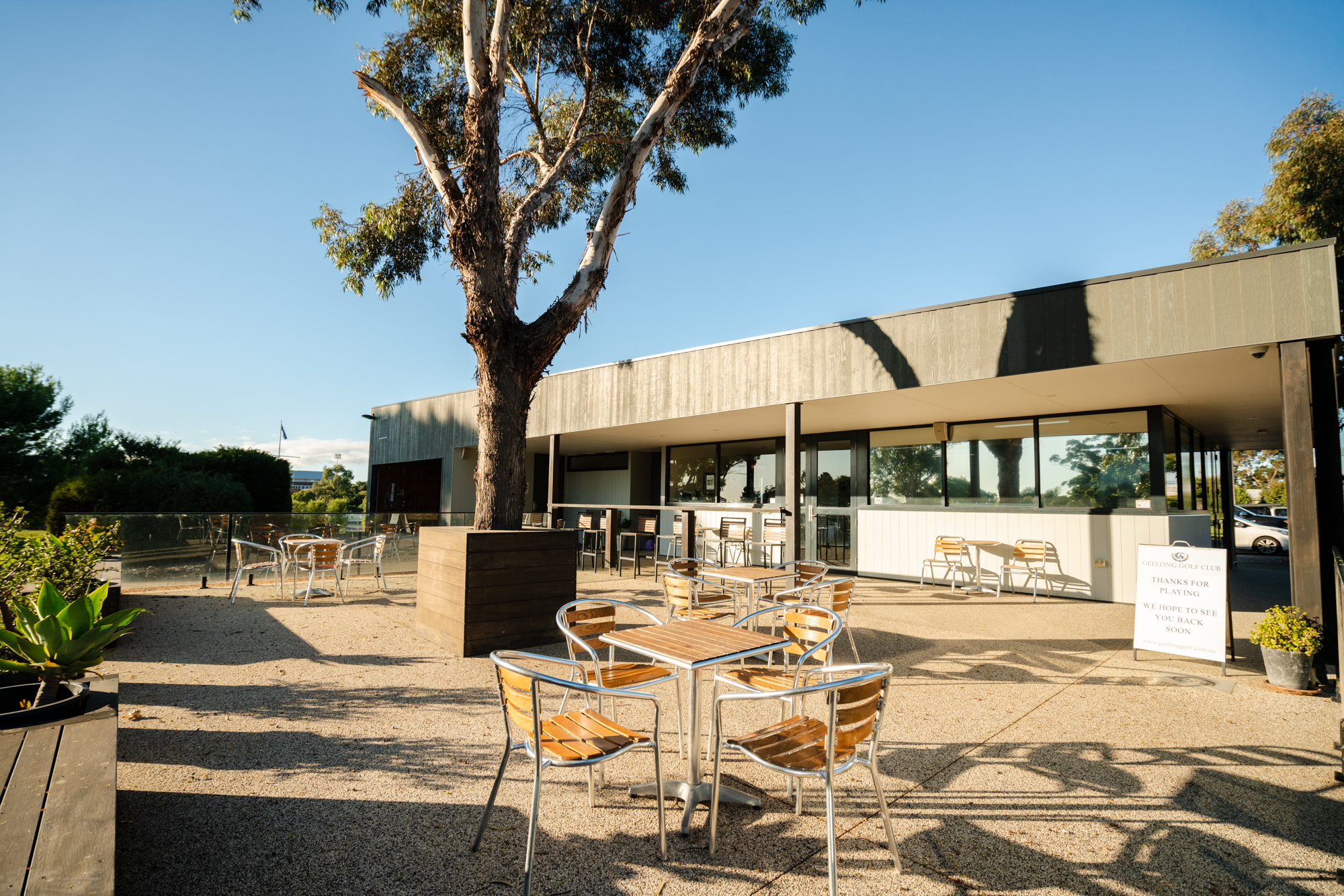 Featured is the outdoor beer garden during the day at Geelong Golf Club. There are outdoor chairs and tables. Behind is the bar.